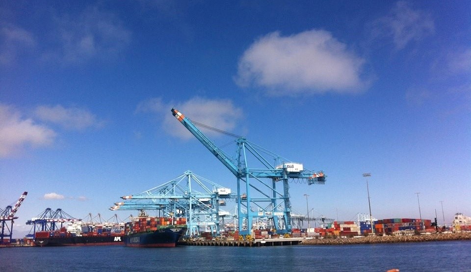 Port scene featuring large cargo ships and blue cranes under a clear sky, showcasing shipping containers and maritime operations.