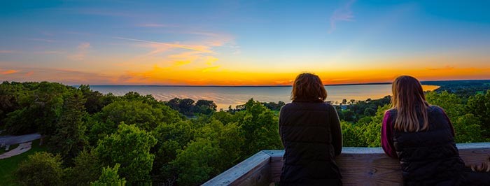 Two women enjoying a vibrant sunset view over a lake, surrounded by lush green trees, while sitting on a wooden balcony.