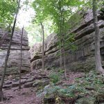 Rock formations surrounded by lush greenery in a forested area, showcasing tall cliffs and rugged terrain. The image captures the natural beauty and geological features of the landscape, ideal for outdoor enthusiasts and nature lovers.