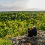 Couple sitting on a rocky outcrop overlooking a lush green forest landscape under a bright sky, enjoying a peaceful moment in nature.
