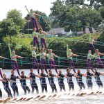A synchronized water skiing performance featuring a team of athletes executing complex formations on the water, showcasing skill and coordination with colorful outfits and multiple tiers of skiers.