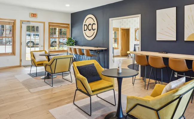 Modern interior of a stylish lounge featuring yellow accent chairs, a sleek black wall with a circular logo, and wooden bar seating. The space is well-lit with natural light, creating a welcoming atmosphere for relaxation and socializing.