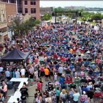 Crowd gathered for an outdoor event in a city plaza, featuring numerous chairs and tents, with local businesses visible in the background. The scene captures the vibrant atmosphere of a community gathering, likely a concert or festival.