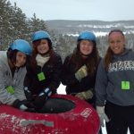 Four young women wearing helmets and winter clothing stand around a red snow tubing inner tube in a snowy landscape. They are smiling and enjoying a winter activity, with trees and a snowy hillside in the background.