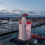 Aerial view of a lighthouse with a red roof and red accents, overlooking a marina filled with boats and a bridge. In the background, large ships are docked at a harbor, with a town and hills visible under a cloudy sky.
