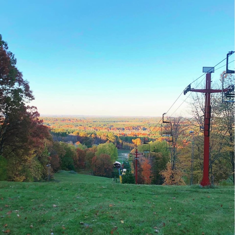 Scenic view from a ski slope overlooking a vibrant autumn landscape with colorful foliage, ski lift chairs in the foreground, and a clear blue sky.