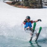 A skier in a blue vest and helmet performs a water ski trick on a snowy slope, creating splashes as they glide over the water. Surrounding trees provide a scenic backdrop, highlighting the thrill of winter sports.