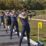 Athletes participating in a biathlon training session, aiming their rifles at targets during a competition on a shooting range, with colorful fall foliage in the background.