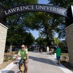 Bicyclist passing through the entrance of Lawrence University, showcasing the iconic archway and vibrant campus atmosphere during an event, with people gathered in the background.