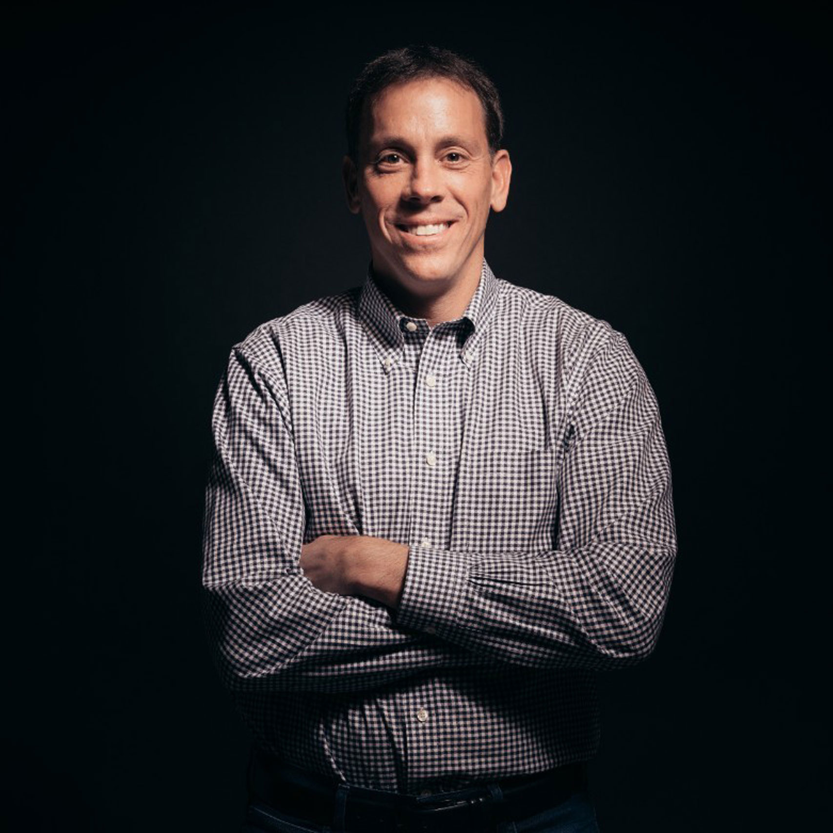 Portrait of a smiling man with crossed arms, wearing a checkered shirt, against a dark background, conveying confidence and professionalism.