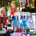 Two women enjoy shopping at a vibrant outdoor market, examining colorful hats and accessories among various handmade crafts. Brightly colored scarves and other items hang in the background, creating a lively atmosphere.