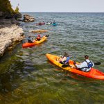 Group of kayakers in orange and red kayaks paddling along a rocky shoreline on a clear lake, surrounded by green trees and a calm blue sky.