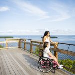 A woman and a girl in a wheelchair enjoy a scenic view from a wooden observation deck overlooking a serene lake and green islands under a clear blue sky.