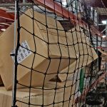 Warehouse scene featuring stacked cardboard boxes secured by a safety net. The image highlights storage organization and safety measures in a commercial setting, showcasing the importance of preventing package falls.