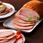 Sliced smoked ham on a platter with fresh herbs and cherry tomatoes, showcasing a juicy and flavorful presentation ideal for sandwiches or holiday meals.