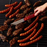 Grilled meats including marinated lamb chops and hot dogs on a barbecue grill, with a hand using a serrated grilling spatula to flip the food. Perfect for summer cookouts and outdoor gatherings.