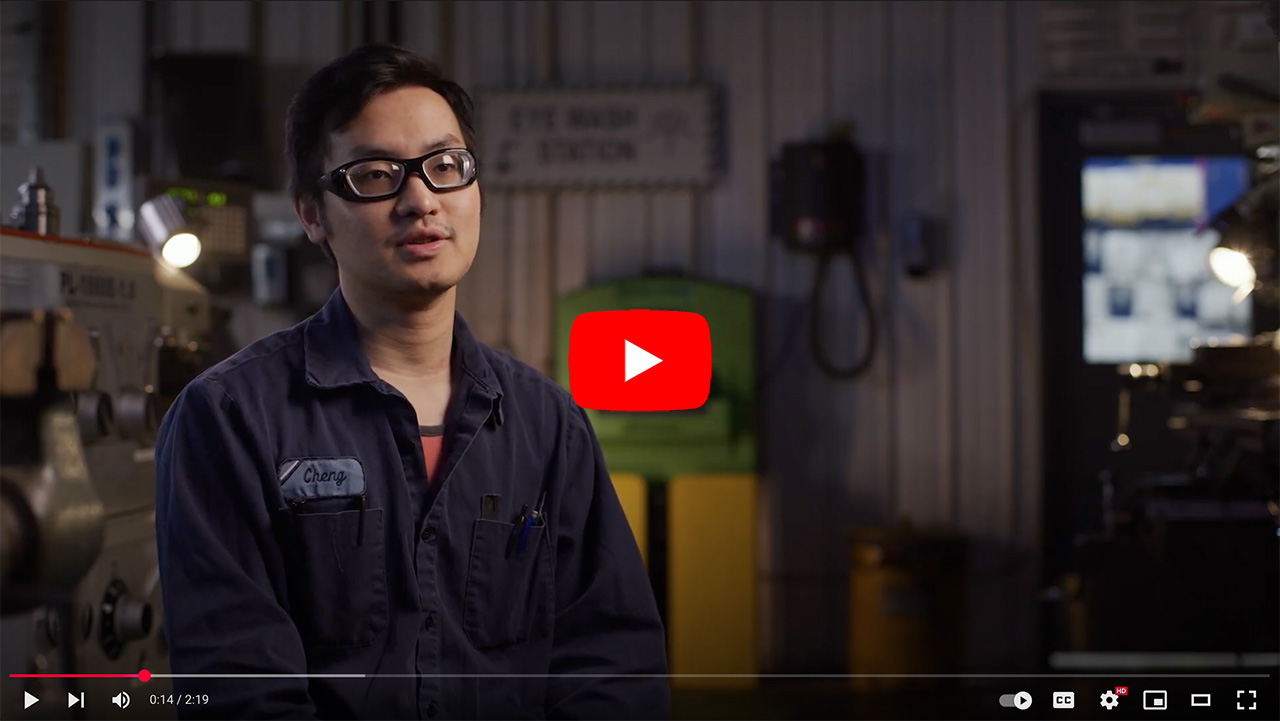 Young man wearing glasses and a work uniform, sitting in a workshop with machinery in the background, discussing his experiences. A YouTube play button is visible, indicating the video format.