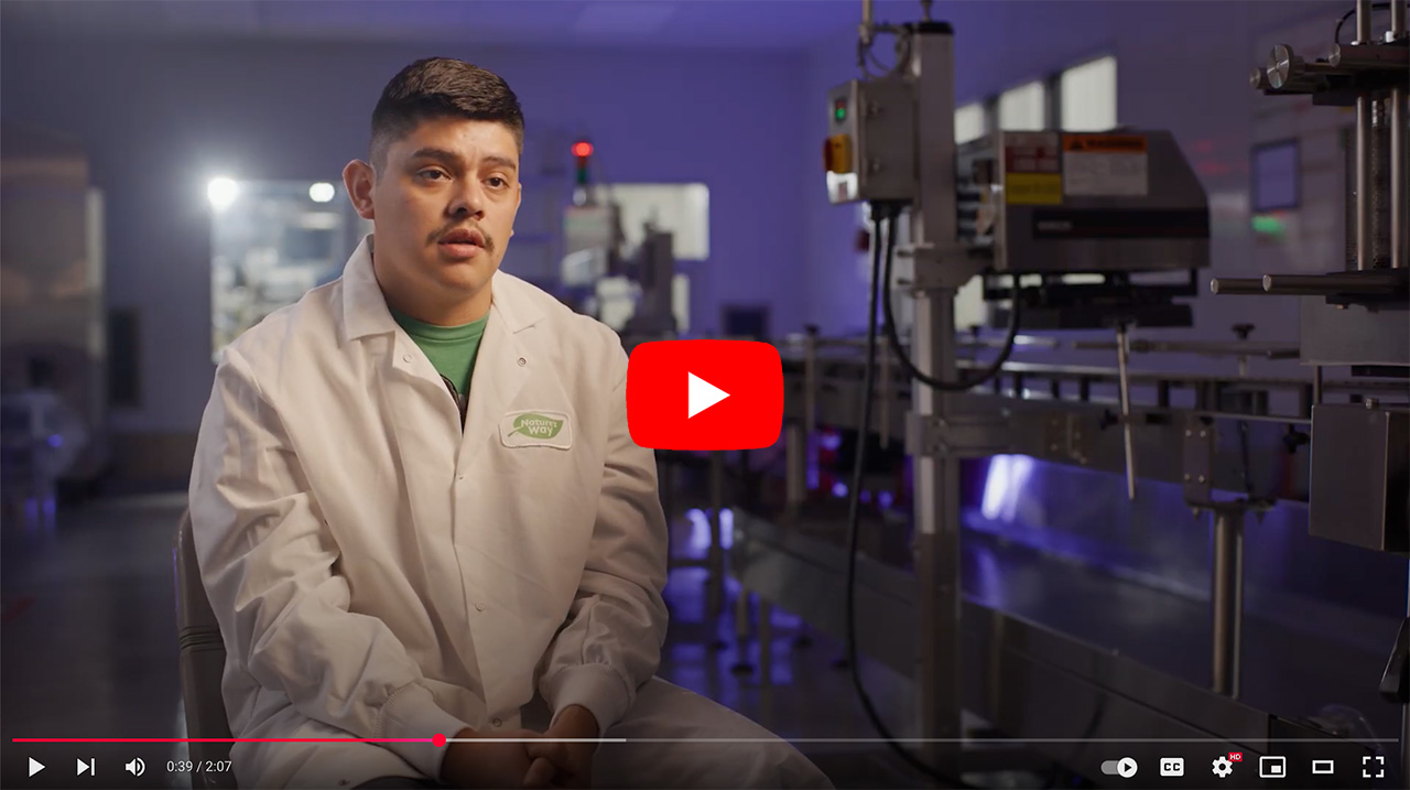 A man in a lab coat sits in a modern production facility, discussing his experiences. The background features machinery and equipment related to manufacturing, highlighting a professional environment focused on quality and innovation.