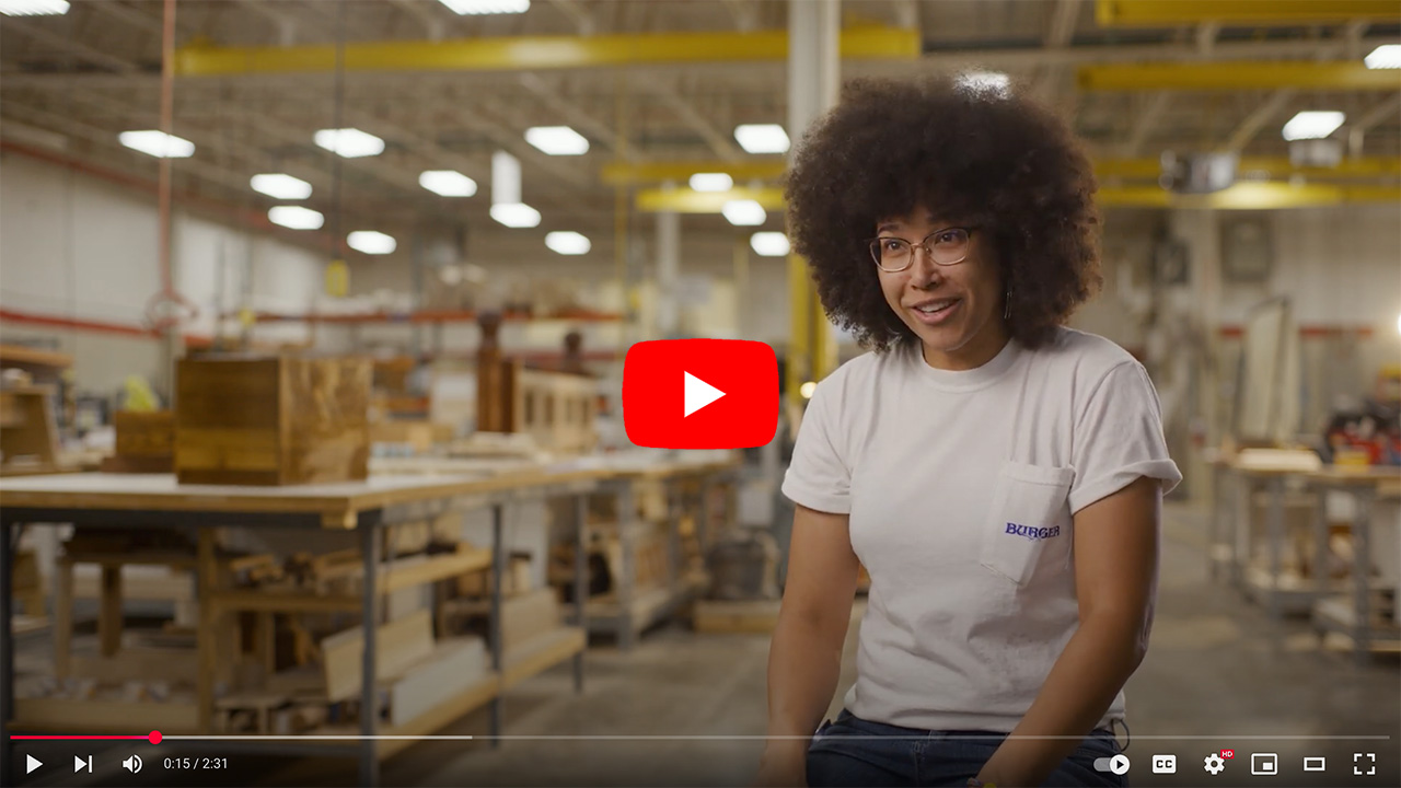Woman with curly hair and glasses sitting in a woodworking shop, smiling while discussing her craft. The background features various woodworking tools and materials, highlighting a creative workspace.