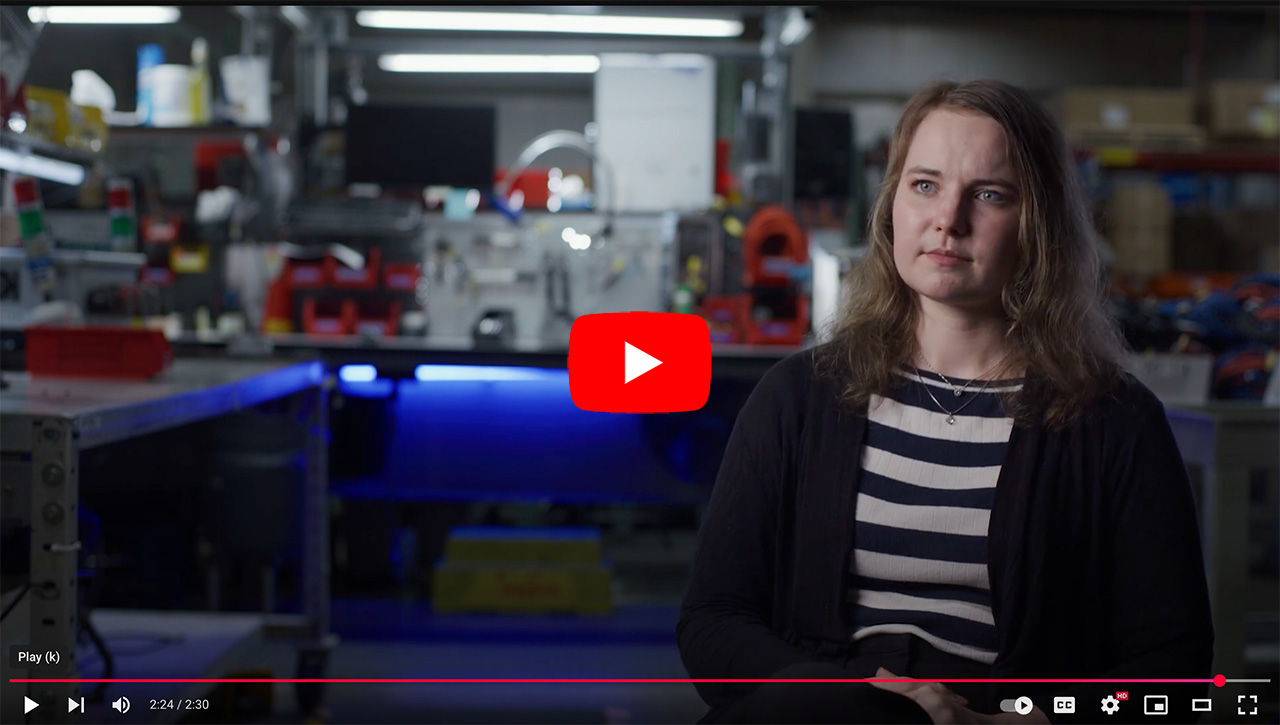 Woman sitting in a workshop setting, discussing technology and innovation, with tools and equipment visible in the background. The scene is well lit, highlighting the organized workspace that reflects a focus on engineering and development.