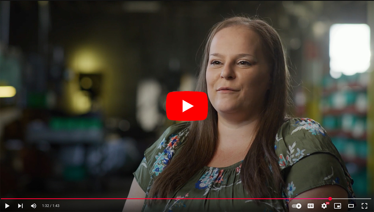 Woman speaking in a casual interview setting, showcasing her insights and experiences. The background features blurred industrial elements, indicating a workplace environment.