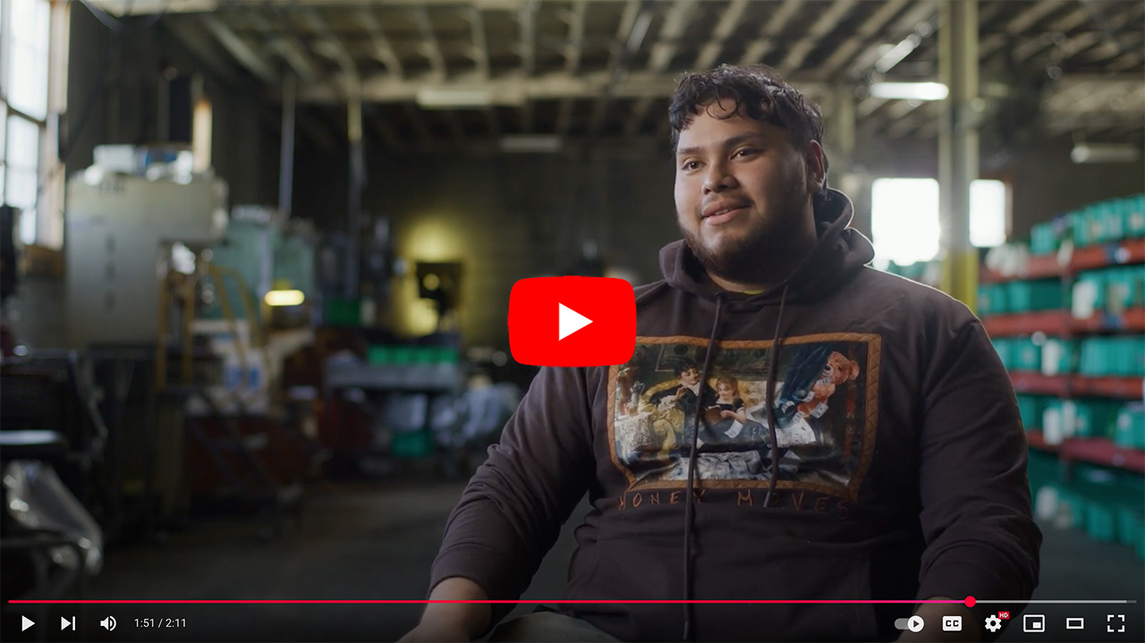 Young man sitting in a casual setting, wearing a graphic hoodie, speaking in an interview context with a blurred background. A YouTube play button is visible, indicating video content.