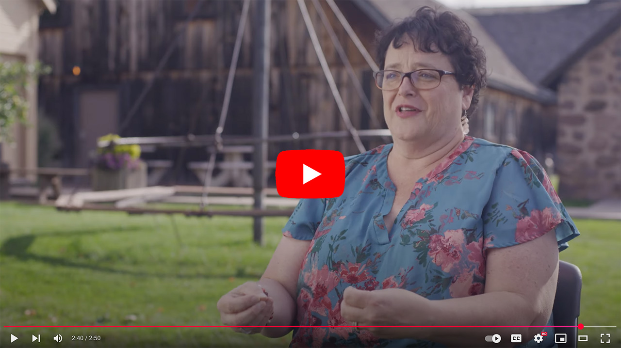 Woman in a floral shirt speaking outdoors in a garden setting, with a rustic barn in the background. The scene captures her engaged in conversation, highlighting themes of storytelling or personal experiences.
