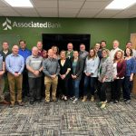 Group photo of Associated Bank employees gathered in a conference room, showcasing teamwork and collaboration. The bank's logo is prominently displayed on the wall, highlighting the corporate environment.
