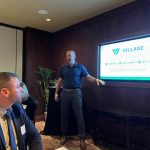 Business presentation in a conference room, featuring a speaker discussing "The Village" focused on companies, investments, and real estate, with an audience member attentively listening.