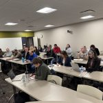 A group of diverse professionals engaged in a training session, seated in a modern classroom with laptops and notebooks on tables. The room features a whiteboard, a clock, and a green accent wall, creating a collaborative learning environment.