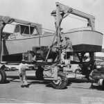 Black and white image of two workers at the Norfolk Naval Shipyard in Portsmouth, Virginia, operating a large boat lift. One worker stands on the ground while the other is seated in the lift, which is hoisting a small boat. The background features industrial buildings and parked cars, capturing a moment in mid-20th century maritime operations.