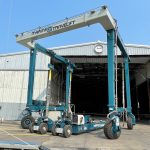 Marine Travelift crane positioned in a boatyard, showcasing its robust structure and wheels, with a clear blue sky and a storage facility in the background. Ideal for lifting and transporting boats.