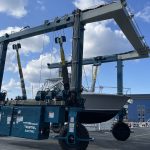 Electric boat lift with a 50 MT capacity, positioned at a marina, lifting a sailboat against a backdrop of blue skies and clouds. The image showcases advanced marine lifting technology for efficient boat handling.