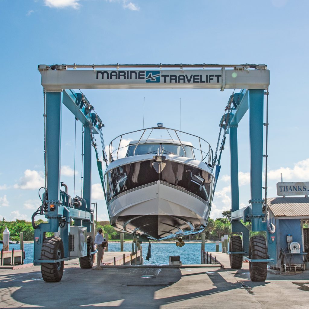 Boat being lifted by a Marine Travelift at a marina, showcasing the lifting equipment and surrounding water. The scene captures the process of boat maintenance and servicing.
