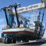 Fishing vessel being lifted at Colonna's Shipyard, showcasing the ship's hull and support equipment against a clear blue sky.