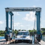 A Marine Travelift lifting a luxury boat at a marina, with workers preparing the vessel for maintenance, surrounded by clear blue skies and greenery.