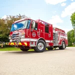 Red fire truck parked on a residential street, showcasing its detailed design, including an American flag on the front and firefighting equipment on the side, surrounded by green trees and a clear blue sky.