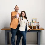 A man and a woman stand in front of a wooden table displaying various beverage cans. The man, dressed in an orange blazer, holds a can while smiling. The woman, wearing a white blazer, also holds a can and smiles at the camera. The background is plain, emphasizing the products they are promoting.