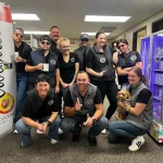 Group photo of a cheerful team wearing matching black vests and casual attire, posing in an office setting with a dog. The image showcases camaraderie and a fun workplace atmosphere, featuring a colorful vending machine in the background.